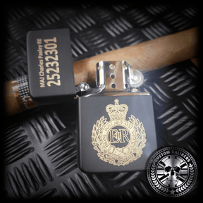 A close up of A matt black lighter with the lid open engraved with the royal engineers crest resting on a cuban cigar